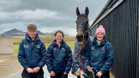 Three people in navy jackets stand beside a racehorse wearing a blanket outside a stable, with rolling hills and a cloudy sky in the background