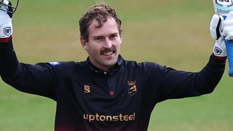 A smiling Ruan de Swardt holds aloft his bat in one hand and his helmet in the other as he celebrates a century against Lancashire