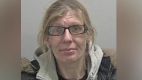 A blonde woman with glasses is wearing a grey hoodie and black coat and looking at the camera against a grey background