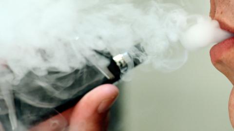 A man is using a black vape. There is smoke coming from his mouth.