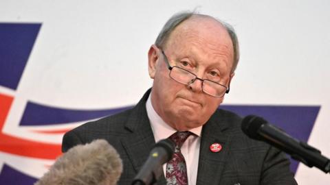 Jim Allister wearing glasses standing behind microphones