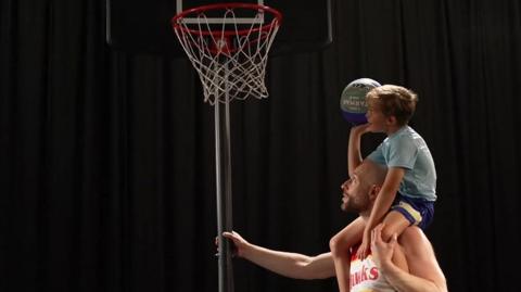 Six-year-old Luther on his dad's shoulders aiming to shoot the basketball through the hoop