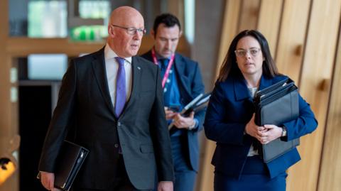 John Swinney, a bald man with glasses, walks to the chamber with Kate Forbes who has long brown hair and is wearing glasses, a blue suit and a white top