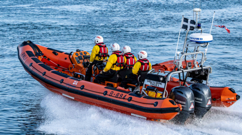Five crew members on board Looe inshore lifeboat, wearing bright yellow waterproof clothing, lifejackets, and white helmets. Their boat is a high-speed, red rigid inflatable, with two outboard motors on the back.