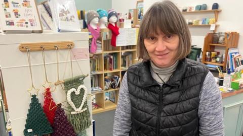 Helen Fallows has short brown hair and is standing in her shop in front of a number of items, like Christmas decorations. She is wearing a black gilet and grey top.