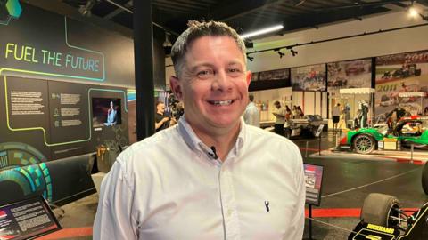A man in a pale blue shirt stands in a room with exhibitions of cars smiling at the camera.
