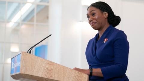 Kemi Badenoch wearing a blue blazer standing on the podium in front of a microphone