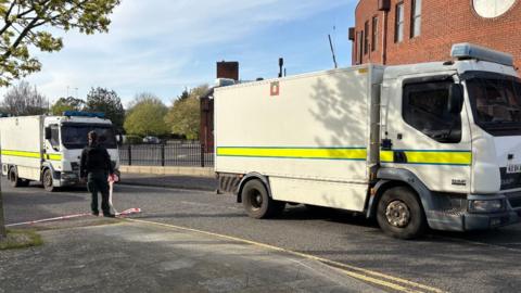 Two large white police trucks parked in front of a red brick building. A police man, dressed all in black, is stood between the trucks, back to camera, holding white and red cordon tape in a residential street. There are trees and railings visible in the background.