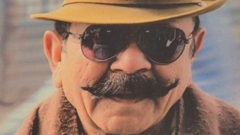 A man with a black moustache wearing a yellow hat and sunglasses. The background behind him is blurred.