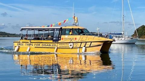 Dolphin III is a small yellow passenger boat, with a roof, and passengers sat in the back. It also has multi-coloured bunting on top, and 'Free Trips for the Disabled' sign-written on the side. It sails through the harbour, a small yacht and the coast in the background.