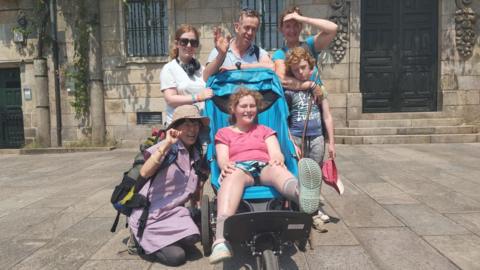 A family in front of an old building. In the middle is a teenage girl in a buggy, surrounded by her mum and dad and a brother and sister.