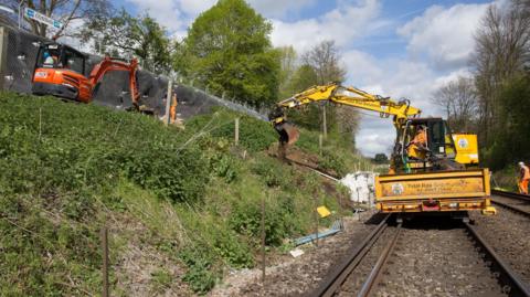 A yellow digger on train tracks and an orange one on embankment working to stabilise the land