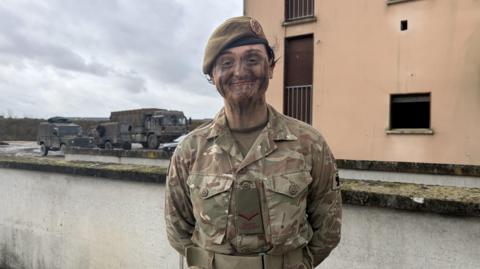 A woman in army combat gear and camouflage face paint stands in front of a wall, she has a green badge saying Duke of Lancaster's. Army vehicles are behind her along with a pink and cream building.