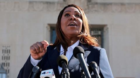 A woman wearing a suit speaks in front of microphones.