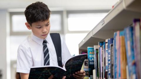 A boy wearing a school uniform stands in a library, reading an open book. He has short dark hear and wears a white shirt with a navy and white striped tie. Shelves filled with books line the right side of the image. There are desktop computers on desks in the background.