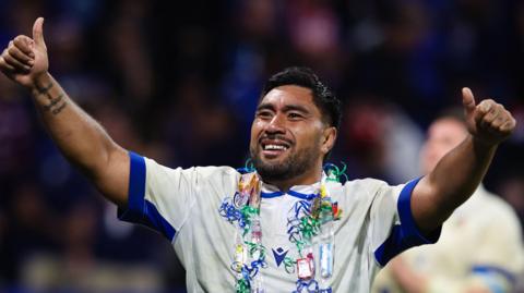Hame Faiva wearing a white Italy kit with thumbs up on both hands, a big smile and daisy-chain style thing around his neck