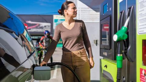 Woman pumping gas. She's wearing a brown long-sleeved t-shirt and camel-coloured trousers. She's squinting in the sun.