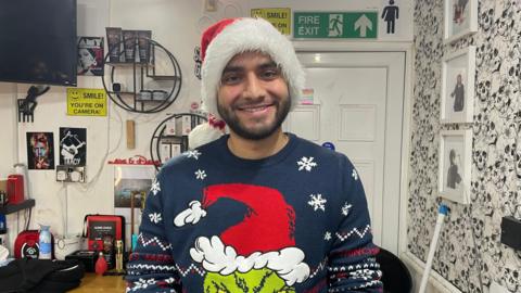 Umar Ali is stood inside the barber shop and is wearing a Grinch Christmas jumper and a red Santa hat. Behind him are pictures of the wall and posters of musicians, amongst various hair products. 