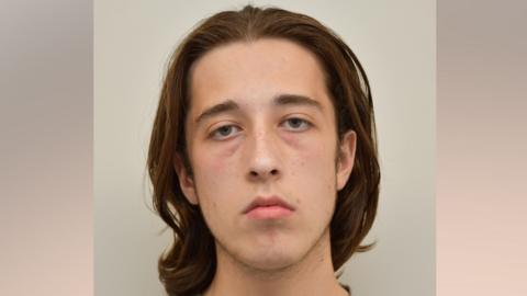 Alfie Coleman has shoulder-length brown hair swept backwards. He looks tired but shows no emotion while pictured in this custody mugshot.