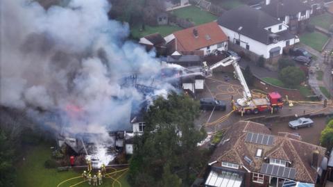 An aerial view of a residential area showing one large house on fire. There are thick plumes of smoke rising from the house on the left. A fire engine is on the driveway up to the house