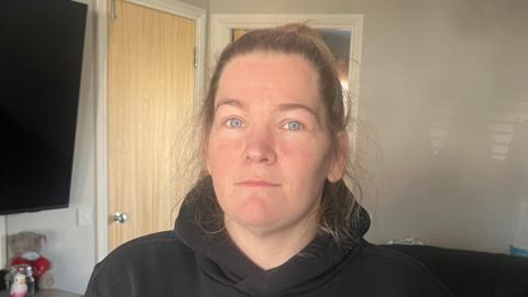 Heather MacMichael, a brown-haired woman wearing a black hoodie, stands in her living room, a grey wall behind her and a TV on the wall to her right.