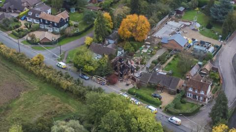 A drone shot of the a destroyed detached house close to other large houses which are untouched. Police tape and emergency vehicles fill the road outside