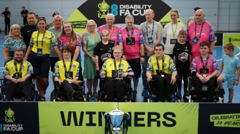 Team shot of Leeds PFC with the trophy standing before them. A yellow and black banner is behind the trophy which reads WINNERS. The players sit in their wheelchairs, while coaches, family and friends stand behind.