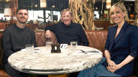 A young man on the left wearing a dark sweatshirt, an older man to his right with red hairt weating a black jumper and a young woman to the right of the photo. She has a blonde bob and wearing a blue blazer with jeans. They are sitting around a round marble-topped table and all looking at the camera and smiling.