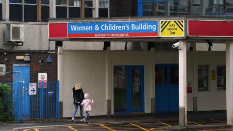 A woman and child walk into a hospital building under a blue sign saying 'Women & Children's Building'