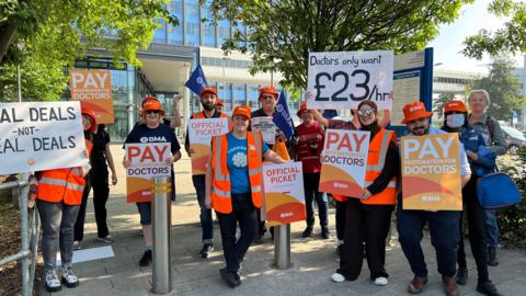 A group of people holding up banners on a doctors strike outside a hospital. Several people are wearing orange hats, hi-vis orange jackets and tops that say BMA. Some of the signs read "Doctors only want £23/hour", "Pay restoration for doctors" and "Official picket".