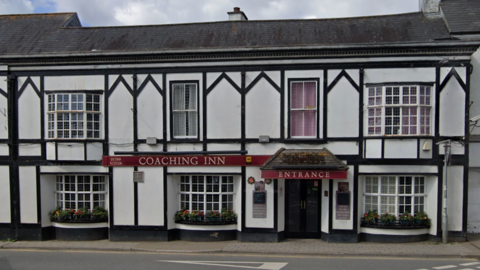 A Google Maps image capturing a white old fashioned building with black panelling, a door with ENTRANCE in red above it, to the left is COACHING INN sign with red background. Cloudy skies behind.