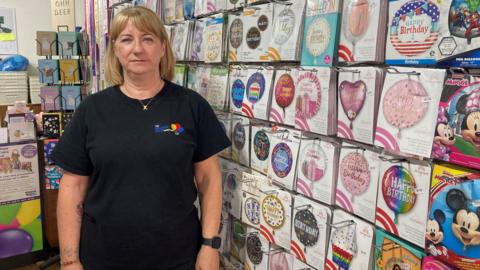 A woman wearing a black t-shirt, stood in a shop next to rows of balloons.