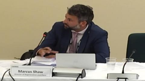 Marcus Shaw appears to be talking into a microphone while sitting at a desk during a council meeting. A sign in front of him includes his name and job title.