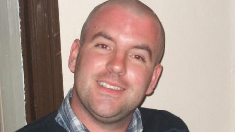 A smiling man wearing a shirt with a dark striped jumper on top