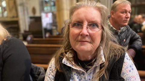 Ms Mooring wears glasses and looks into the camera from where she is sitting in the pew.