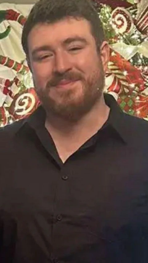 A picture of Zak Hughes. He is smiling at the camera. He is wearing a black, button up shirt and has dark hair and facial hair.