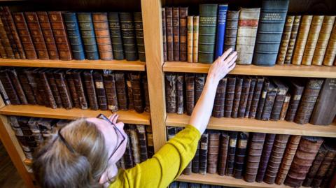 A woman wearing a yellow top and glasses reaches for a book from a bookshelf.