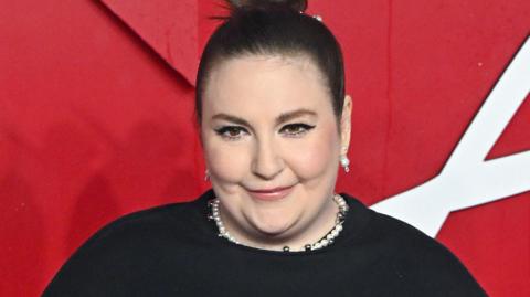 Lena Dunham, in a black dress, smiling at the camera