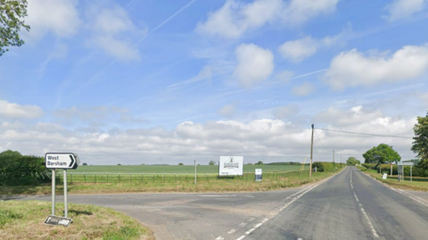 Street view of a straight stretch of the B1105 on a sunny day. There is an intersection either side of the road and a white road sign, pointing right, towards 'West Barsham'. 