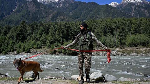 A man in camouflage stands beside a river with a dog on a lead and mountain in the background
