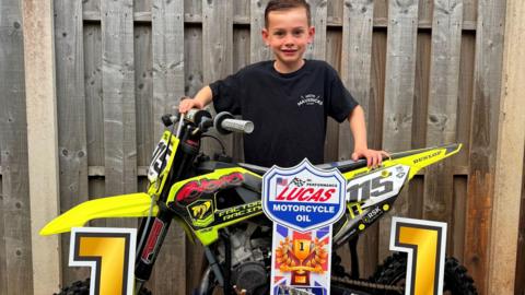 A young boy in a black t-shirt smiles sweetly as he stands behind a yellow motorbike