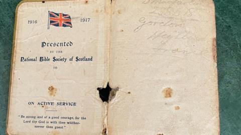The inside of a bible with script on the left-hand side, a hole between the two open pages towards the bottom and some faint writing 'D MacFarlane Gordon Highlanders France' on the right-hand side page.