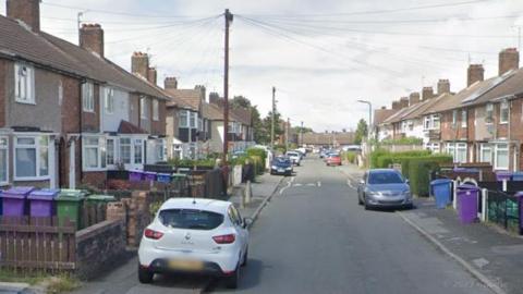A road of terraced houses with a number of cars parked half on the pavement.