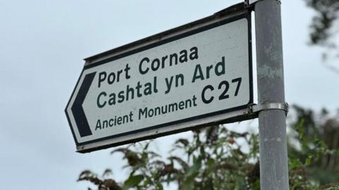 A white arrow road sign pointing left reads: Port Cornaa, Castal yn Ard, in front of a grey sky and green foliage.