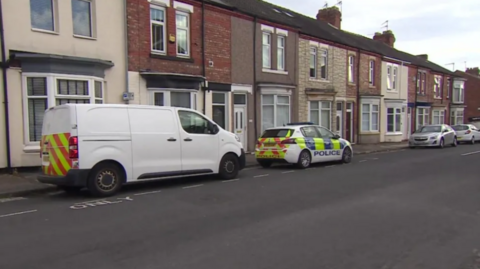 View of a police car and a white van parked on a street of two-storey houses.