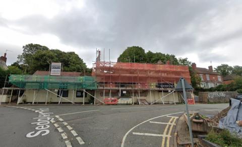 A long building which has scaffolding and netting over it. It is on the corner of a road. 
