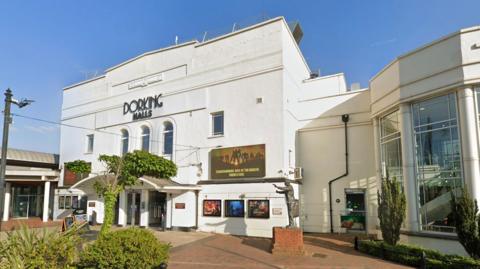 The outside of a theatre. The building is white with film posters on the outside and black letters that spell 'Dorking Halls'. 