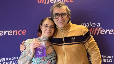 A man and a woman are stood with their arms round each other in front of a purple banner that says 'BBC Radio Sheffield' and 'BBC Make A Difference' 