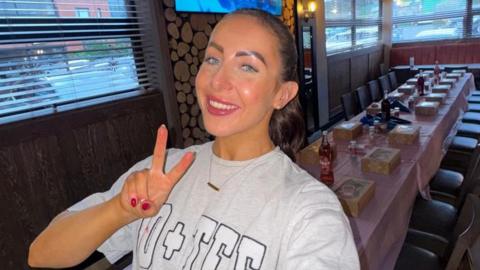 A selfie of Ashlin McCourt wearing a grey jersey sweatshirt with her dark hair slicked back. She is smiling and making a peace sign while working as a waitress with a long table seen behind her