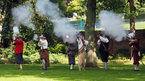 A group of six men in period clothing fire muskets in the air. The weather is sunny and then men are stood beside a large tree. 